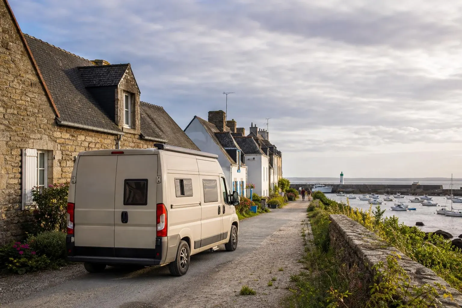 Fourgon aménagé blanc vu de trois-quarts arrière, garé dans une ruelle de pierre d'un village portuaire breton, un phare et des bateaux au mouillage visibles au loin, deux passants sur le chemin